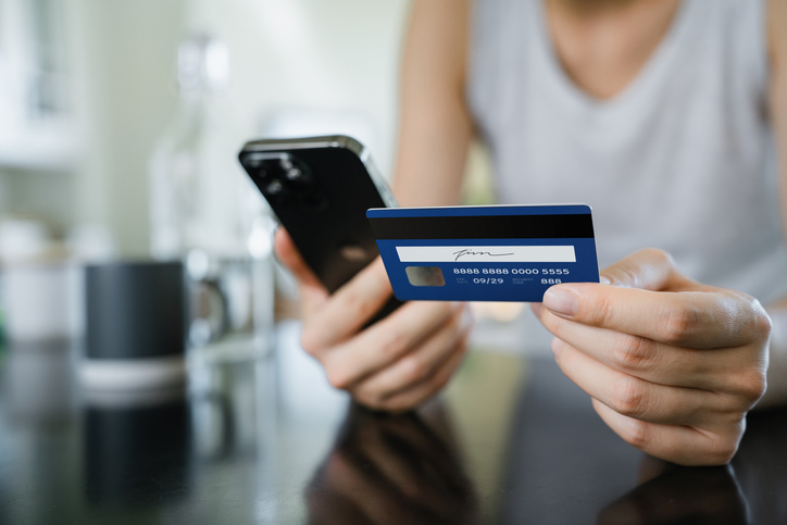 Close-up shot of young Asian woman settling a payment with smartphone and credit card over the kitchen counter at home. Shopping online, paying bills via digital banking. Contactless payment and e-commerce concept. Lifestyle and technology. Smart banking